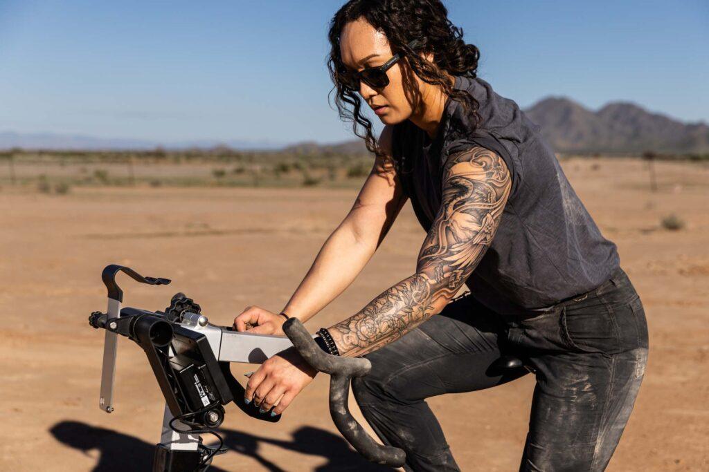 Tatuerad kvinna som använder cyckelmaskin ute i öken.