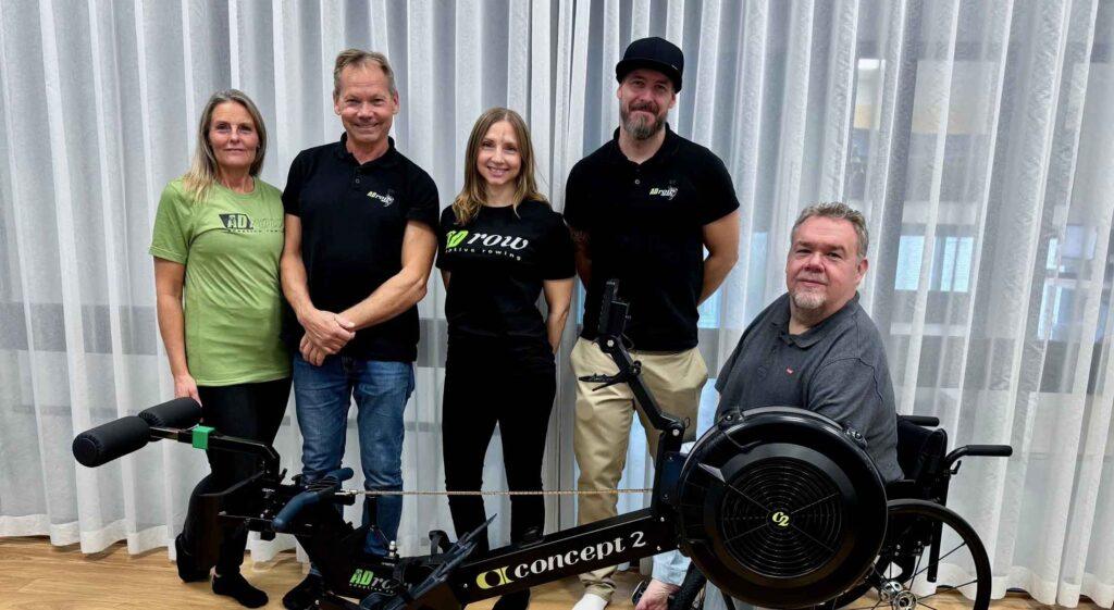 En leende grupp som står vända mot kameran framför en rowerg som är specialanpassad för inkludering